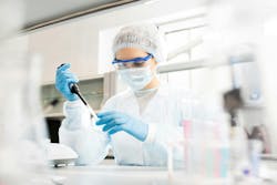 Serious concentrated female microbiologist in sterile clothing and safety goggles sitting at table and dropping reagent in petri dish while doing research in laboratory